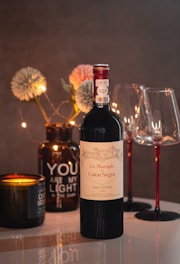 A bottle of red wine labeled 'Le Marquis de Calon Ségur' stands prominently on a glossy surface. Surrounding it are three wine glasses with red stems, two floral arrangements with soft pink and white flowers, and two candles, one of which is lit, creating a warm ambient light. The setting suggests an elegant and cozy atmosphere.