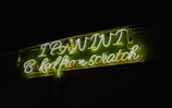 Bright neon signage displaying the text 'I PAINT Baked from scratch' in glowing green and white letters. The surrounding background is dark, enhancing the vividness of the neon lights.