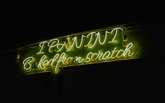 Bright neon signage displaying the text 'I PAINT Baked from scratch' in glowing green and white letters. The surrounding background is dark, enhancing the vividness of the neon lights.