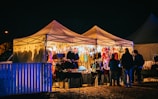 A nighttime scene featuring a brightly lit market stall filled with various colorful items such as clothing and accessories. Several people are gathered around, browsing the selections, with the warm glow of the lights contrasting against the dark night sky.