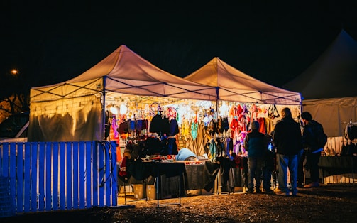 A nighttime scene featuring a brightly lit market stall filled with various colorful items such as clothing and accessories. Several people are gathered around, browsing the selections, with the warm glow of the lights contrasting against the dark night sky.