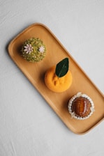 A wooden tray holds three artfully crafted desserts. The dessert on the left is covered in green crushed nuts with small pink flower decorations. The middle dessert is round, orange, and textured, topped with a single dark green leaf. On the right, a glossy caramelized dome is surrounded by white cream or frosting petals.