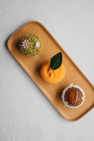 A wooden tray holds three artfully crafted desserts. The dessert on the left is covered in green crushed nuts with small pink flower decorations. The middle dessert is round, orange, and textured, topped with a single dark green leaf. On the right, a glossy caramelized dome is surrounded by white cream or frosting petals.