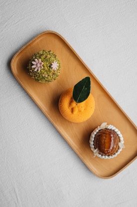 A wooden tray holds three artfully crafted desserts. The dessert on the left is covered in green crushed nuts with small pink flower decorations. The middle dessert is round, orange, and textured, topped with a single dark green leaf. On the right, a glossy caramelized dome is surrounded by white cream or frosting petals.
