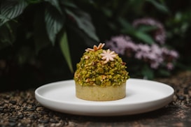 A dessert topped with chopped pistachios and decorated with small edible flowers is placed on a white plate. The background features green foliage and blurred purple flowers, creating a natural and lush setting.