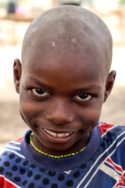 A young child with a shaved head is smiling openly, displaying an expression of joy. The background is softly blurred, focusing attention on the child's face. The child is wearing a colorful shirt with a pattern of stripes and polka dots, along with a beaded necklace.