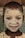 A child with a painted face smiles at the camera. The face is covered with a pale, clay-like substance, highlighting the eyes and mouth. The child is wearing a patterned garment, and their hair is short.