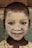 A child with a painted face smiles at the camera. The face is covered with a pale, clay-like substance, highlighting the eyes and mouth. The child is wearing a patterned garment, and their hair is short.