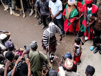 A group of people encircle a central performer dressed in a striking, striped costume. Some individuals are dressed in vibrant traditional clothing, while others have cameras and phones raised, capturing the scene. The setting appears to be a street or open space, with onlookers gathered closely.