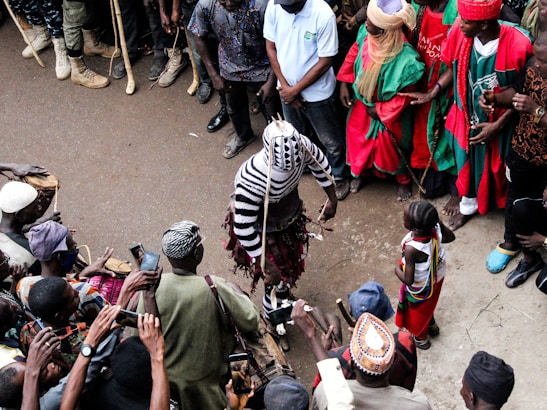 A group of people encircle a central performer dressed in a striking, striped costume. Some individuals are dressed in vibrant traditional clothing, while others have cameras and phones raised, capturing the scene. The setting appears to be a street or open space, with onlookers gathered closely.