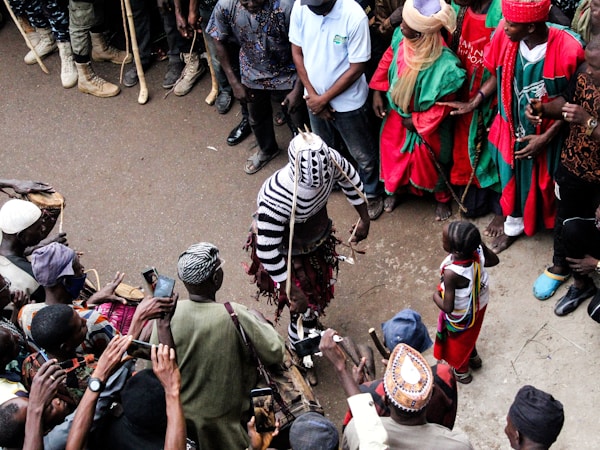A group of people encircle a central performer dressed in a striking, striped costume. Some individuals are dressed in vibrant traditional clothing, while others have cameras and phones raised, capturing the scene. The setting appears to be a street or open space, with onlookers gathered closely.