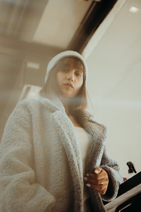 A person wearing a cozy, textured coat and a beanie stands indoors. The setting has a warm, muted lighting, adding a soft, introspective atmosphere.