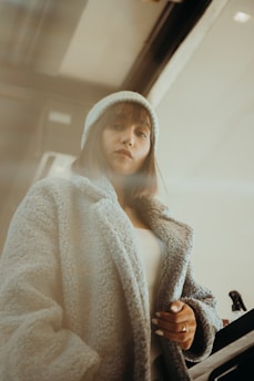 A person wearing a cozy, textured coat and a beanie stands indoors. The setting has a warm, muted lighting, adding a soft, introspective atmosphere.