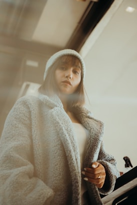 A person wearing a cozy, textured coat and a beanie stands indoors. The setting has a warm, muted lighting, adding a soft, introspective atmosphere.