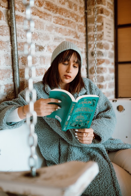 A person wearing a cozy blue coat and a matching beanie is sitting on a swing indoors, engrossed in reading a book with a turquoise cover. The background consists of a rustic brick wall and metallic chains.
