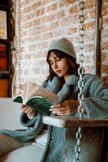 A person is reading a book while seated on a swing indoors. They are wearing a cozy blue or teal coat and a matching beanie. The background is a wall with exposed bricks, giving the scene a rustic and warm ambiance.