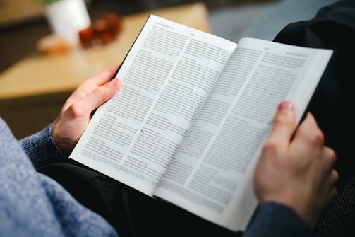A person wearing a blue sweater is holding an open book and reading, with visible text and a focus on the hands and the book. The background is slightly blurred, giving prominence to the book and hands.