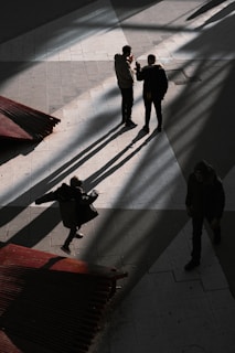 Three people stand on a tiled ground, their long shadows cast dramatically due to the angle of the light. The sun creates sharp contrasts between light and dark. A figure appears to be in mid-motion, perhaps dancing or playfully moving, while the others seem engaged in conversation. Red structures on the left add a pop of color against the otherwise monochromatic setting.