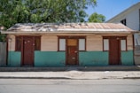 A modest, single-story house with a corrugated metal roof and wooden walls painted half brown and half teal. There are three doors and three windows with shutters painted in dark brown. The house stands on a simple concrete foundation with a dirt-surfaced foreground. Lush, green trees with a clear blue sky are visible in the background.