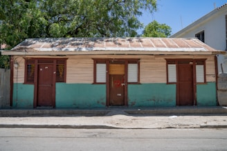 A modest, single-story house with a corrugated metal roof and wooden walls painted half brown and half teal. There are three doors and three windows with shutters painted in dark brown. The house stands on a simple concrete foundation with a dirt-surfaced foreground. Lush, green trees with a clear blue sky are visible in the background.