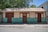 A modest, single-story house with a corrugated metal roof and wooden walls painted half brown and half teal. There are three doors and three windows with shutters painted in dark brown. The house stands on a simple concrete foundation with a dirt-surfaced foreground. Lush, green trees with a clear blue sky are visible in the background.