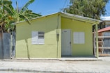 A small house painted in light green with a white door and two white shuttered windows. The house has a simple design and a metal roof, situated on a concrete surface next to a dirt road. Banana trees and other greenery are visible beside and behind the house, while a wooden and metal fence partially surrounds it.