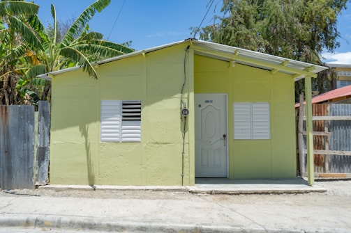 A small house painted in light green with a white door and two white shuttered windows. The house has a simple design and a metal roof, situated on a concrete surface next to a dirt road. Banana trees and other greenery are visible beside and behind the house, while a wooden and metal fence partially surrounds it.