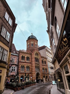 A historic cityscape with a prominent brick building featuring ornate arches and a domed tower topped with a cross. The building is surrounded by narrow streets lined with other architectural structures. People are visible entering or leaving the building, and there are outdoor seating arrangements with umbrellas.