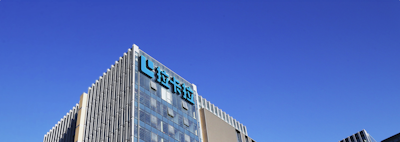 A modern office building with glass walls reflecting the clear blue sky. The structure has a prominent logo on the upper part, consisting of stylized blue letters.