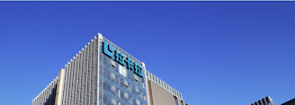 A modern office building with glass walls reflecting the clear blue sky. The structure has a prominent logo on the upper part, consisting of stylized blue letters.