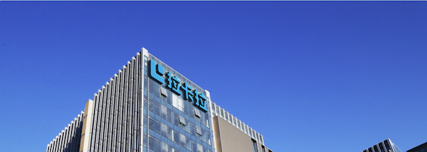 A modern office building with glass walls reflecting the clear blue sky. The structure has a prominent logo on the upper part, consisting of stylized blue letters.