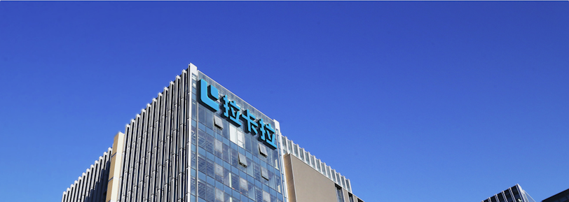 A modern office building with glass walls reflecting the clear blue sky. The structure has a prominent logo on the upper part, consisting of stylized blue letters.