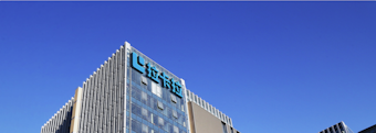 A modern office building with glass walls reflecting the clear blue sky. The structure has a prominent logo on the upper part, consisting of stylized blue letters.