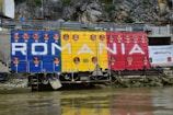 A wall mural along a riverbank displays the word 'ROMANIA' against a backdrop of large horizontal bands in blue, yellow, and red, resembling the Romanian flag. Multiple bronze busts are attached to the mural above and below the letters. There are tires attached to the structure, and a banner with the text 'Dacia Renaste' is seen to the side. The setting includes rock formations and a railing above the mural.