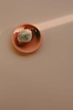 A single green and orange mandarin with leaves sits on a peach-colored plate with soft sunlight casting a shadow.