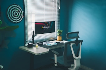 A modern and tidy workspace with a dartboard on the teal wall, lit by natural light through blinds. A computer monitor on the desk displays a colorful screen. The desk hosts a potted plant, a closed notebook, a candle, and some tech gadgets. An ergonomic office chair is positioned nearby.