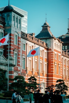 A historic red-brick building with ornate architecture, featuring large windows and intricate detailing. Two Japanese flags are prominently displayed in the foreground. A group of people is seen walking, indicating a lively atmosphere. The building is set against a clear blue sky, and there is a modern high-rise partially visible in the background.
