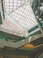 An indoor architectural scene featuring a large atrium with a glass ceiling allowing natural light to flood the space. Multiple levels of walkways and escalators are visible with a few people walking. The design is modern, incorporating clean lines and geometric shapes.