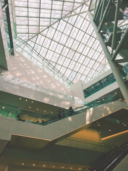 An indoor architectural scene featuring a large atrium with a glass ceiling allowing natural light to flood the space. Multiple levels of walkways and escalators are visible with a few people walking. The design is modern, incorporating clean lines and geometric shapes.