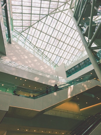 An indoor architectural scene featuring a large atrium with a glass ceiling allowing natural light to flood the space. Multiple levels of walkways and escalators are visible with a few people walking. The design is modern, incorporating clean lines and geometric shapes.
