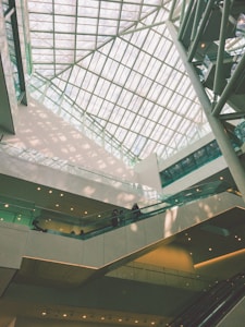 An indoor architectural scene featuring a large atrium with a glass ceiling allowing natural light to flood the space. Multiple levels of walkways and escalators are visible with a few people walking. The design is modern, incorporating clean lines and geometric shapes.