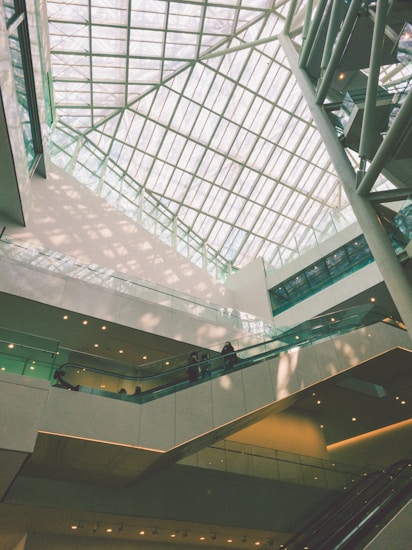 An indoor architectural scene featuring a large atrium with a glass ceiling allowing natural light to flood the space. Multiple levels of walkways and escalators are visible with a few people walking. The design is modern, incorporating clean lines and geometric shapes.