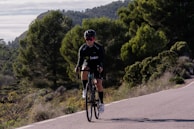 A cyclist in athletic gear is riding a bicycle on a road surrounded by lush greenery and hills. The person is wearing a helmet and sunglasses, and appears to be focused on the ride. The road curves slightly and there are dense trees in the background, suggesting a rural or mountainous setting.