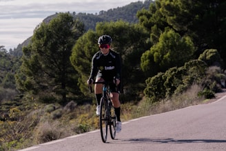 A cyclist in athletic gear is riding a bicycle on a road surrounded by lush greenery and hills. The person is wearing a helmet and sunglasses, and appears to be focused on the ride. The road curves slightly and there are dense trees in the background, suggesting a rural or mountainous setting.