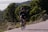 A cyclist in athletic gear is riding a bicycle on a road surrounded by lush greenery and hills. The person is wearing a helmet and sunglasses, and appears to be focused on the ride. The road curves slightly and there are dense trees in the background, suggesting a rural or mountainous setting.