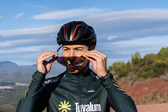 A cyclist wearing a helmet and sunglasses adjusts the gear on a scenic outdoor trail. The background shows hills and a partly cloudy sky, indicating a sunny day. The attire is a long-sleeved sports jersey with visible branding.