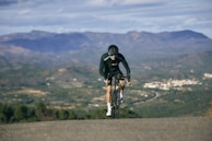 A cyclist in black cycling gear rides up a slight incline, set against a backdrop of expansive mountains and a sprawling valley. The landscape is dotted with small clusters of buildings and roads. The overall atmosphere is one of determination, with the cyclist focused and pushing forward under a partly cloudy sky.