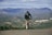 A cyclist in black cycling gear rides up a slight incline, set against a backdrop of expansive mountains and a sprawling valley. The landscape is dotted with small clusters of buildings and roads. The overall atmosphere is one of determination, with the cyclist focused and pushing forward under a partly cloudy sky.