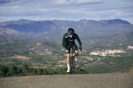 A cyclist in black cycling gear rides up a slight incline, set against a backdrop of expansive mountains and a sprawling valley. The landscape is dotted with small clusters of buildings and roads. The overall atmosphere is one of determination, with the cyclist focused and pushing forward under a partly cloudy sky.