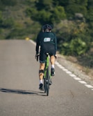 A cyclist in full gear rides along a smooth, empty road surrounded by greenery. The rider is wearing a dark uniform and helmet, showcasing an athletic posture. The bicycle has a green frame, and shadows are cast on the road surface.